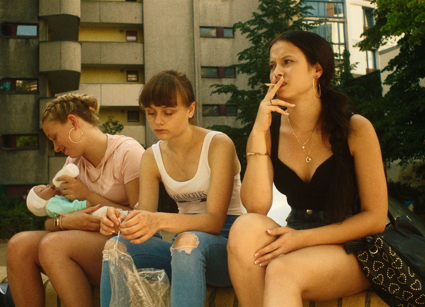 Trois adolescentes assises sur un muret. Des immeubles se dressent derrière. L'une des jeunes femmes fume, une autre regarde ses mains, la dernière nourrit une poupée.